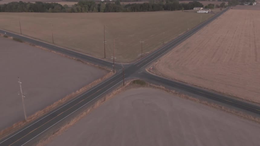 An aerial shot slowly tilts down over a desolate crossroads cutting through vast dry farmland at dusk