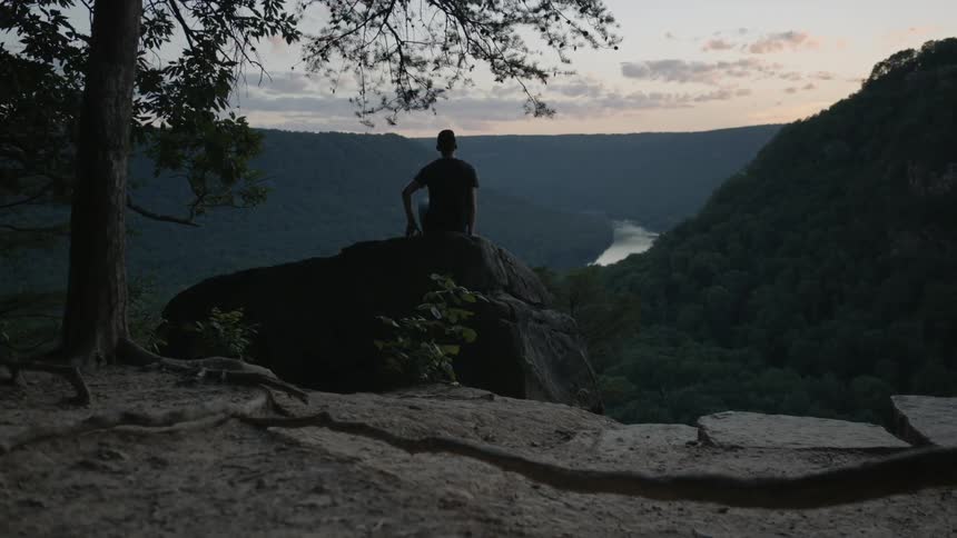 A man walks and sits on a boulder overlooking a valley in the wilderness