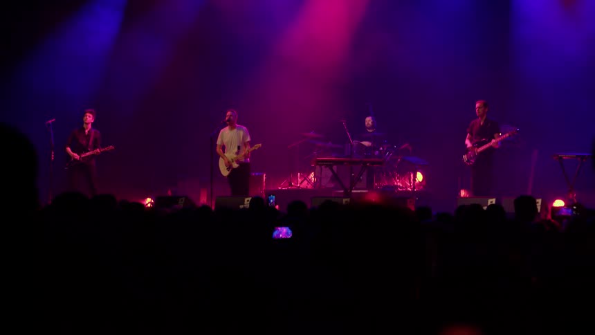 A live band performs on stage under vibrant purple and red lighting in front of a cheering crowd