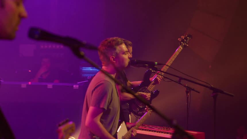 Three musicians perform onstage with guitars and microphones under purple and orange lighting