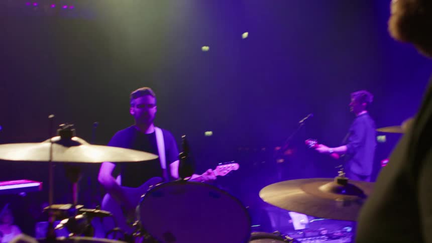 A band performs on stage under purple lighting with the drummer in the foreground and guitarists playing in the background