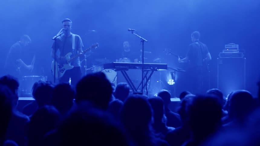 A man singing and playing guitar at a rock concert for a large crowd alongside other musicians playing percussion drums and bass