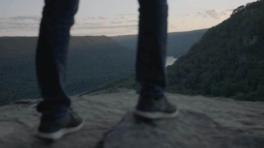 Person walking to the edge of a cliff to sightsee mountains and a river