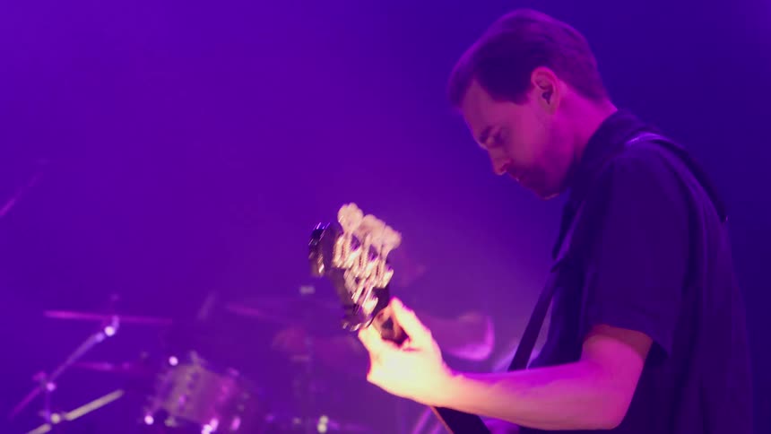 A guitarist and drummer perform live on stage under vibrant purple lighting