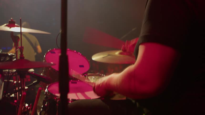 A drummer performs onstage under bright lighting singing into a microphone positioned near the drum kit