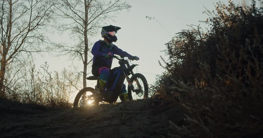 A motocross rider pauses at the top of a dirt trail in a wooded area at sunset