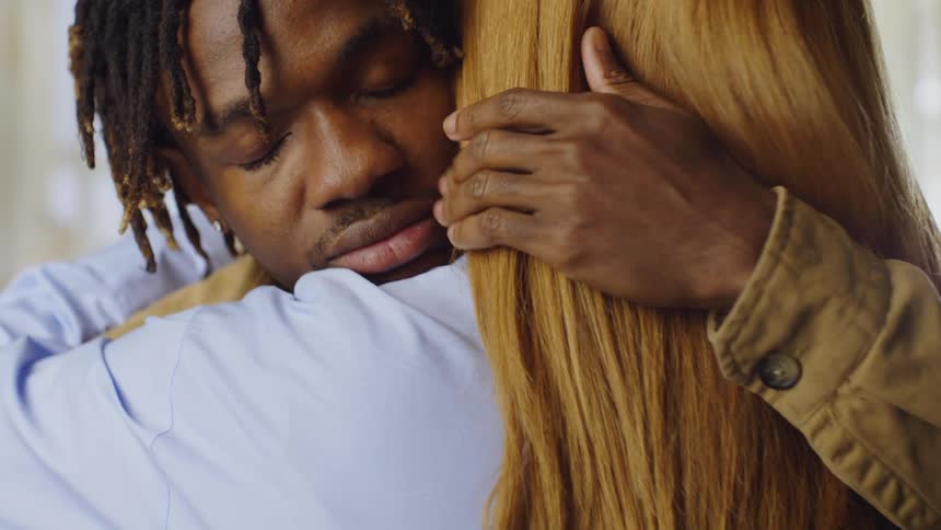 A young black man hugs a blonde woman with long hair gently rubbing his hand on her back