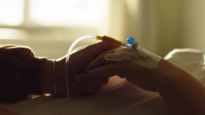 Pair of hands clasped together one of which is a patient lying in a hospital bed