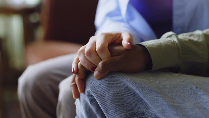 A young lady sits beside a black man whose leg is shaking nervously she gently places her hand on his resting atop his knee in an effort to calm him down as they sit together on a couch in the living room