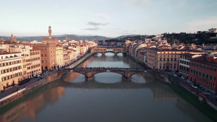 A bridge that passes over a canal that is full of people on it and cars passing in the streets on the sides