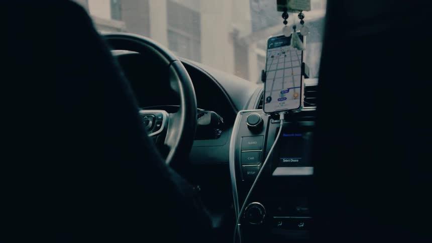 A car interior shows a steering wheel and dashboard with a smartphone mounted on the air vent displaying a navigation map