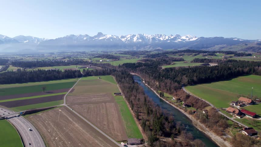 A river flows through a rural landscape with fields forests and scattered houses set against a backdrop of snowcapped mountains