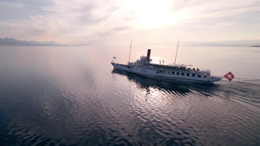A vintage paddle steamer flying a swiss flag serenely cruises across a vast calm lake at sunset with mountains silhouetted on the horizon
