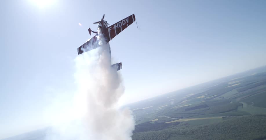 A stunt plane performs a vertical climb with smoke trailing behind it over a vast rural landscape