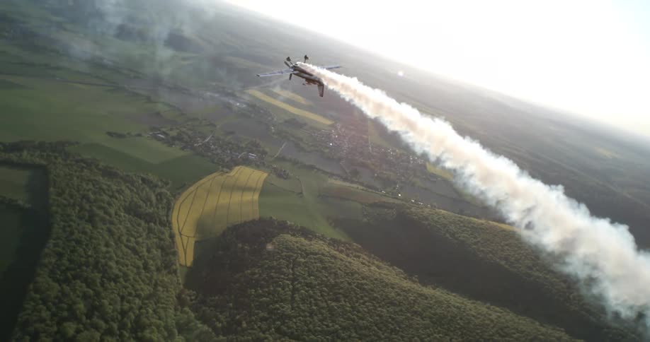An aircraft flies low over patchy farmland and forest trailing a thick plume of white smoke