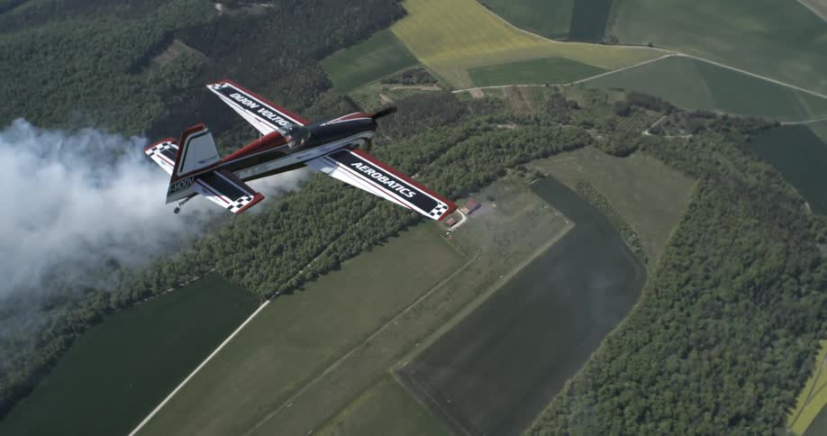 Stunt plane performing aerial maneuver over vast green fields with smoke trailing behind