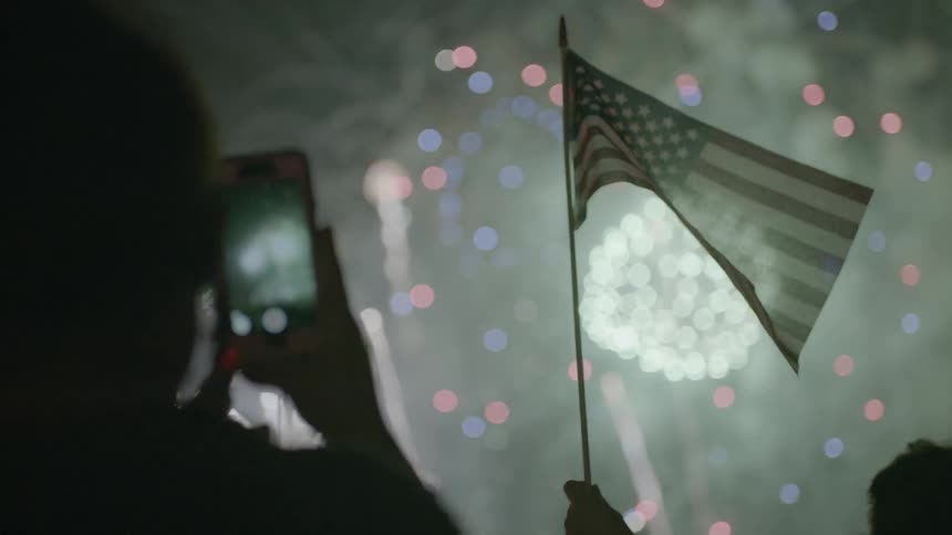 An american flag is waved as fireworks explode overhead