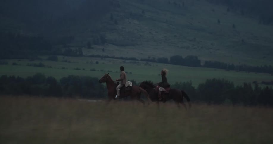 Two people ride horses across a wide field beneath misty mountains at dusk