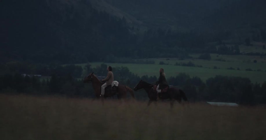 Two people riding horses through a field with mountains in the background