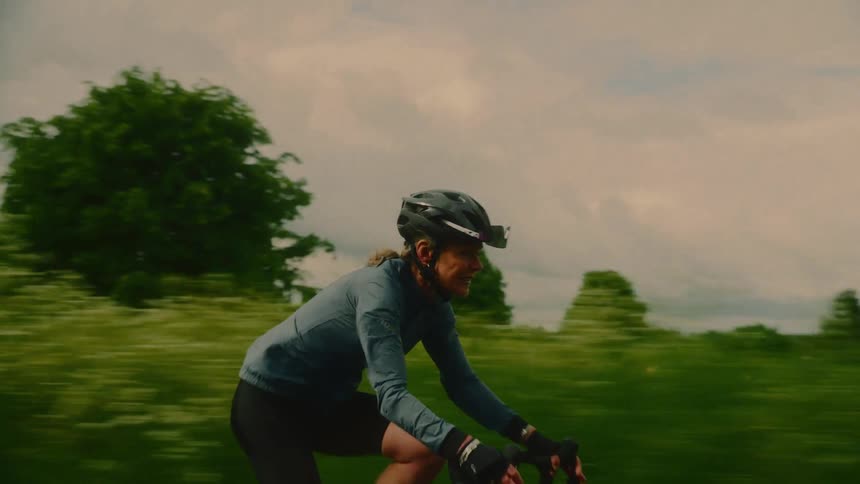 A woman wearing the helmet and is riding fast on a bicycle and behind her there are various vegetations in a field of grass and trees