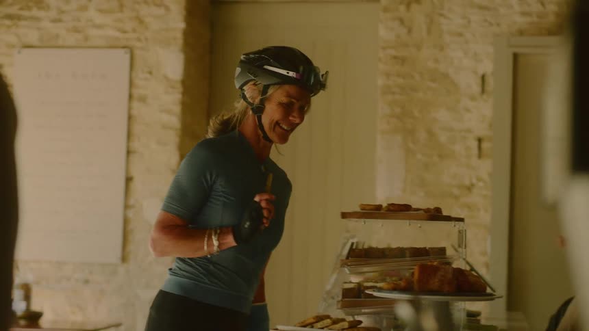 A female cyclist wearing her helmet stands at a buffet telling the server what she wants on her plate as another woman walks by and they greet each other at midday