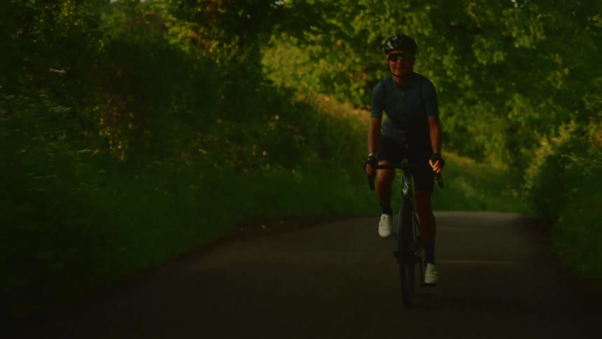 A cyclist in a helmet and sunglasses rides a bicycle towards the camera on a narrow tree lined path