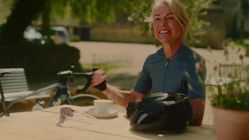 Female cyclist smiles and touches her hair while sitting at a table outdoors during daytime