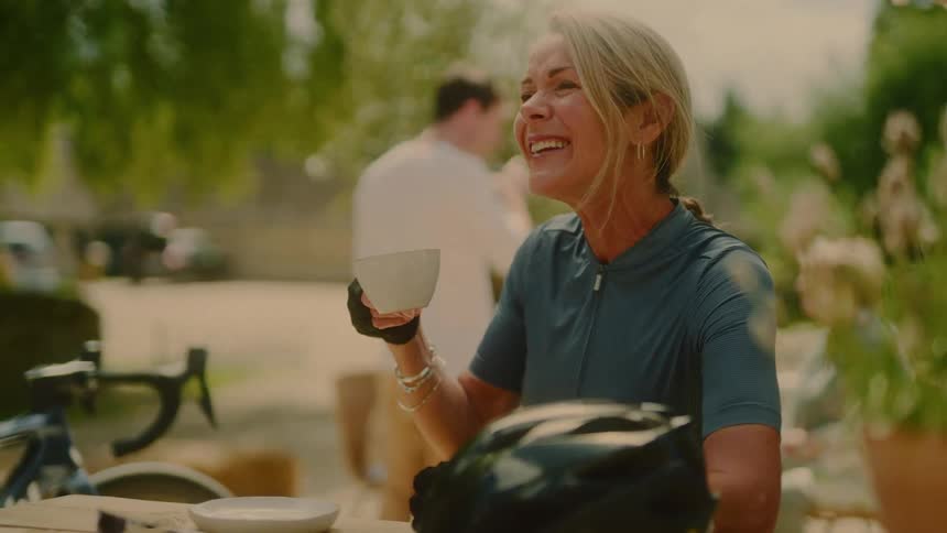 A woman cyclist sits and laughs during a conversation at midday enjoying a cup of coffee her helmet rests on the table and her bike is parked beside her