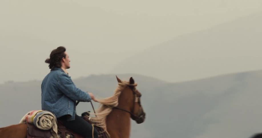 Two people ride horses across an open field with a mountain backdrop