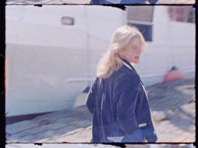 A young woman walks through a boatyard at midday wearing a blue suit does a 360degree turn with a smile and then continues walking