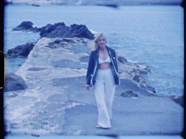 A woman poses on a rocky pier by the sea smiling at the camera