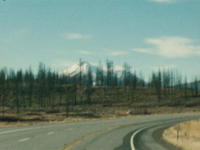 Vehicles drive along a winding road with a snowcapped mountain in the distance