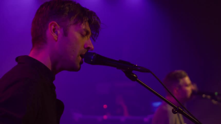 A male guitarist plays the guitar and sings into the microphone while performing with his band during a musical concert