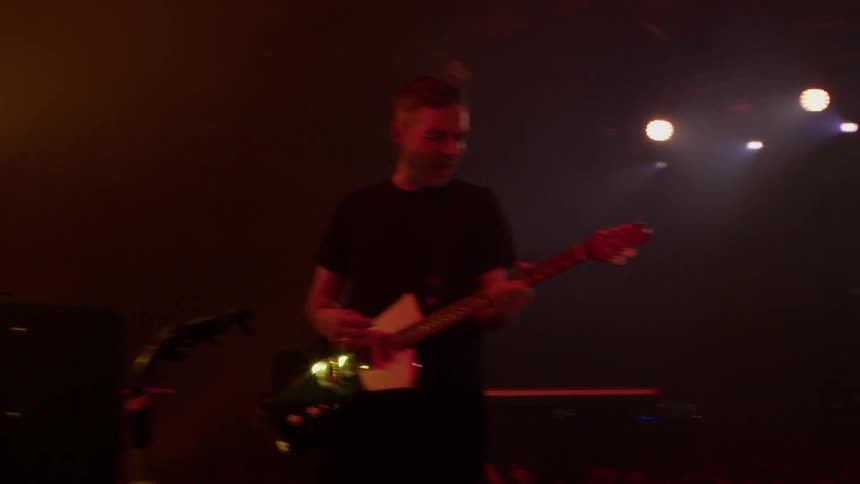 Energetic guitarists perform on a dimly lit stage illuminated by warm hazy spotlights during a live concert
