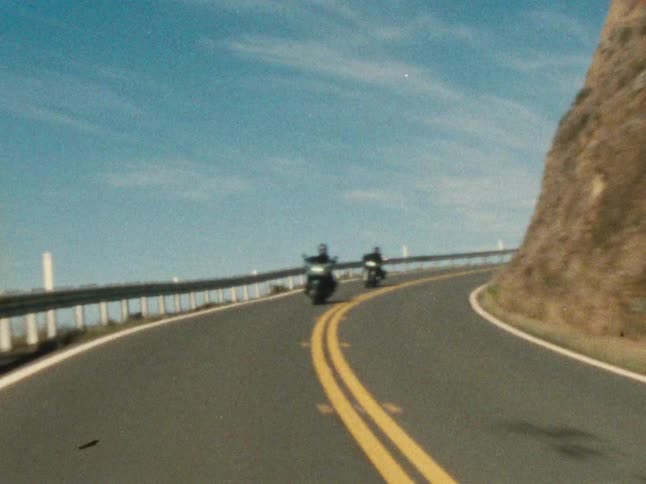 Two motorcyclists ride on a highway curving around the base of a mountain on a cloudy day