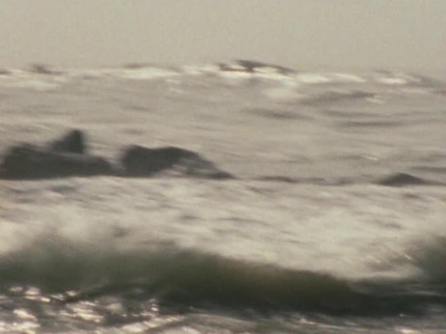 Waves roll past a barrier of rocks near the shore on an overcast day