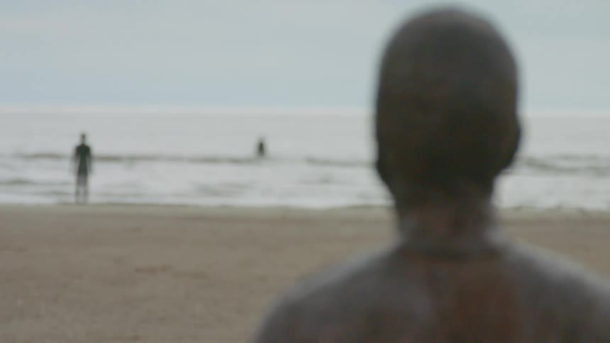 A statue of a person and two people standing on a beach and in the ocean