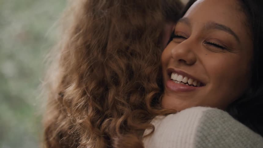 Two young ladies hug each other with smiles on their faces while indoors at midday