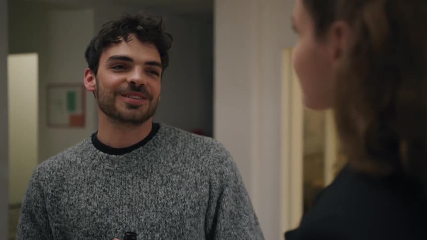 A man wearing a gray sweater is holding a bottle and when he is talking to a woman in an indoor setting