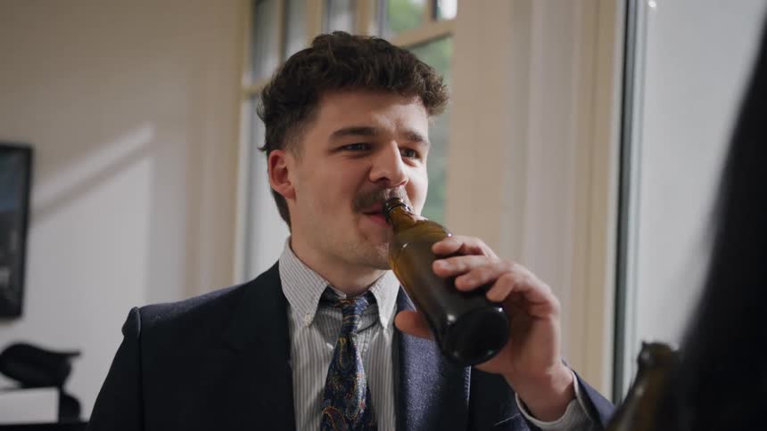 A man in a suit smiles and drinks from a bottle while enjoying a casual conversation indoors