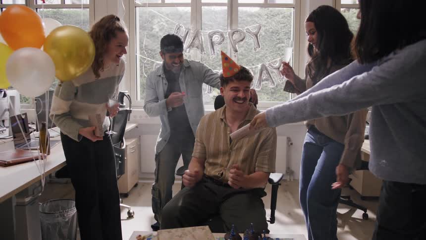 Coworkers celebrate a birthday with dancing and laughter around a seated man in a party hat