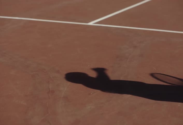 A persons shadow is visible on the floor of a tennis court as they bounce a tennis ball before striking it with a racket on a sunny day