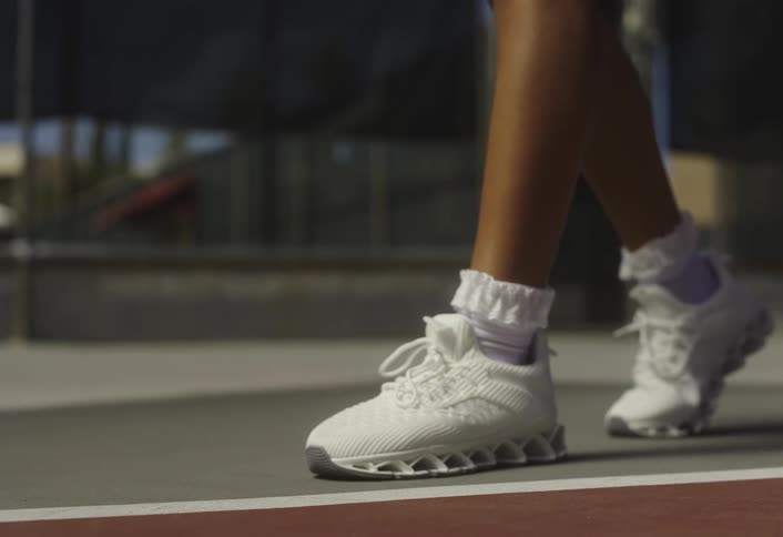 A womans feet wearing white sneakers stand on a tennis court as she bounces a tennis ball on the court at midday