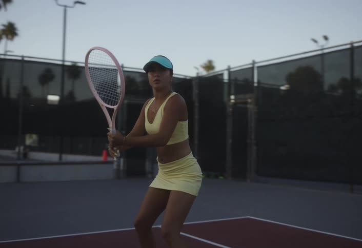 A young lady wearing a yellow top skirt and blue hat strikes the tennis ball on the court during a competitive game