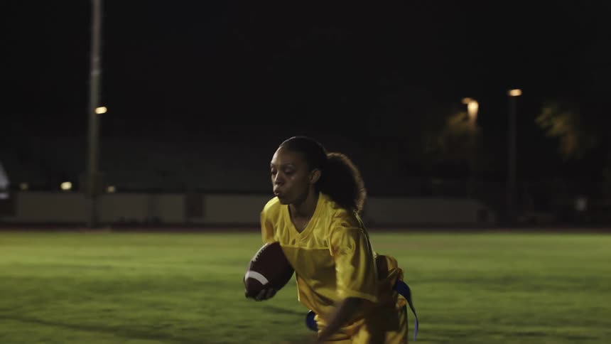 A flag football player in a yellow jersey stretches forward with the ball as defenders close in under stadium lights