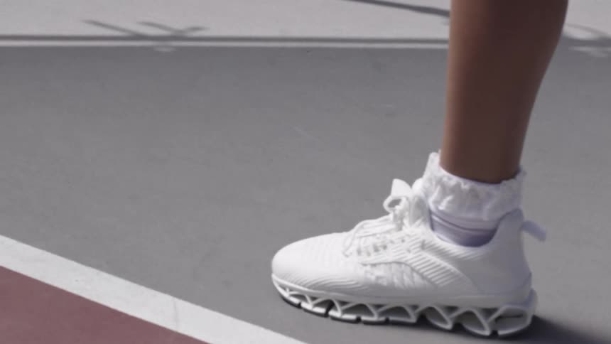 A ball bounces straight up and down on a tennis court beside the foot of a tennis player wearing white sneakers with air pocketed soles and frilly white socks on a bright day