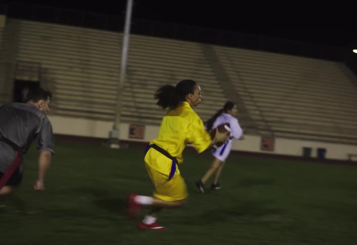 An american football match featuring both men and women takes place at night on a pitch with the ball passed to a woman in a yellow jersey as she runs toward the goal