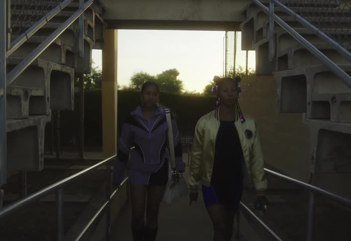 A camera follows two stylish young women as they walk from underneath stadium bleachers out to a landing overlooking a running track and field