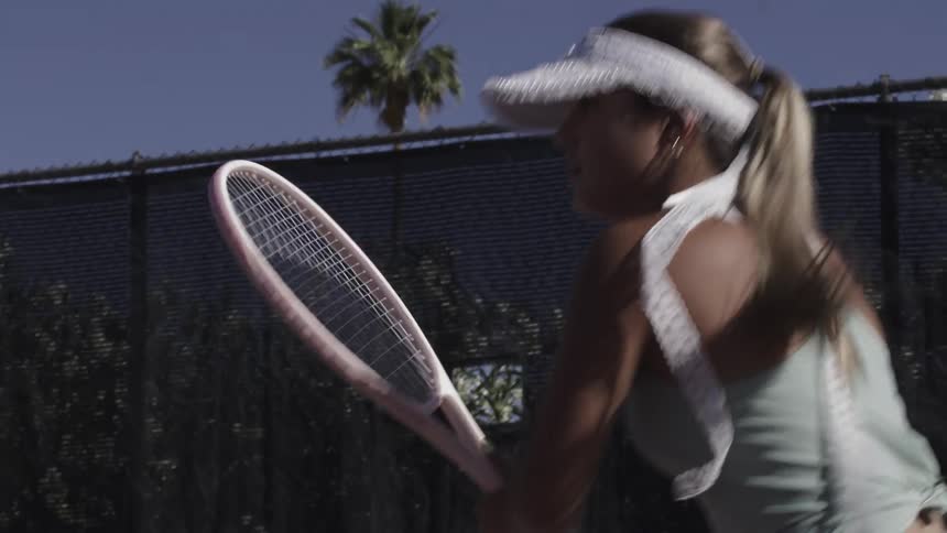 A woman wearing a white visor swings a tennis racket on a court with a fence and palm trees in the background