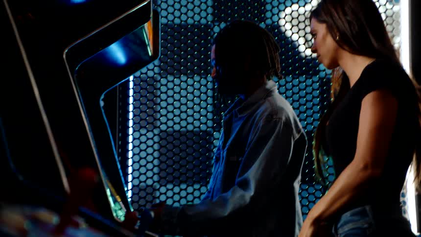 A black man plays an arcade game while a woman stands beside him